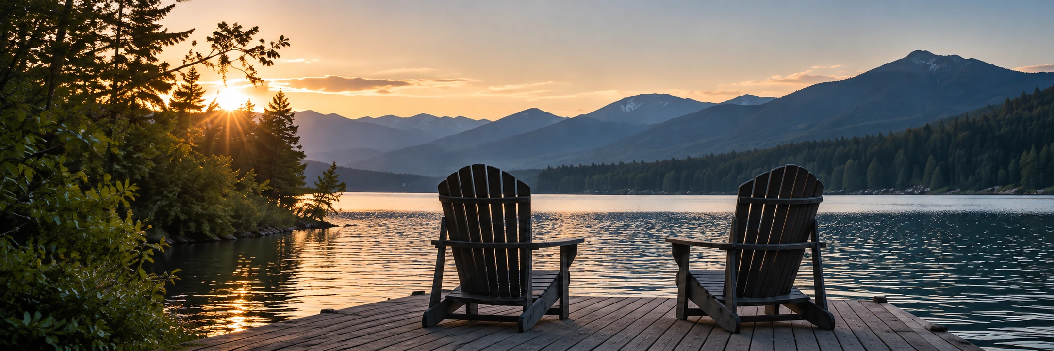 Two wooden chairs on a dock, facing a mountain lake at sunset